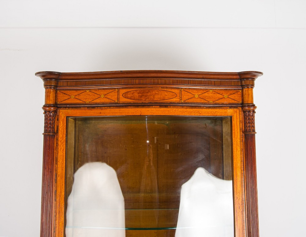 A tall Edwardian satinwood display cabinet featuring a curved glazed door, glass shelves, fluted corner columns, and geometric marquetry inlay. View III