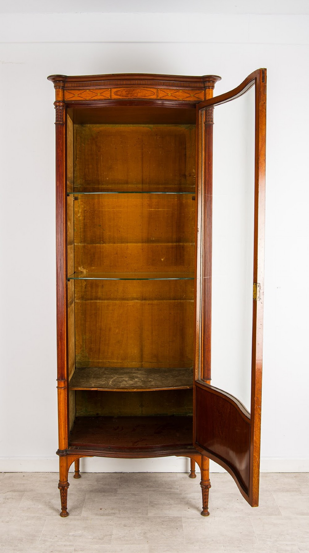 A tall Edwardian satinwood display cabinet featuring a curved glazed door, glass shelves, fluted corner columns, and geometric marquetry inlay. View IV