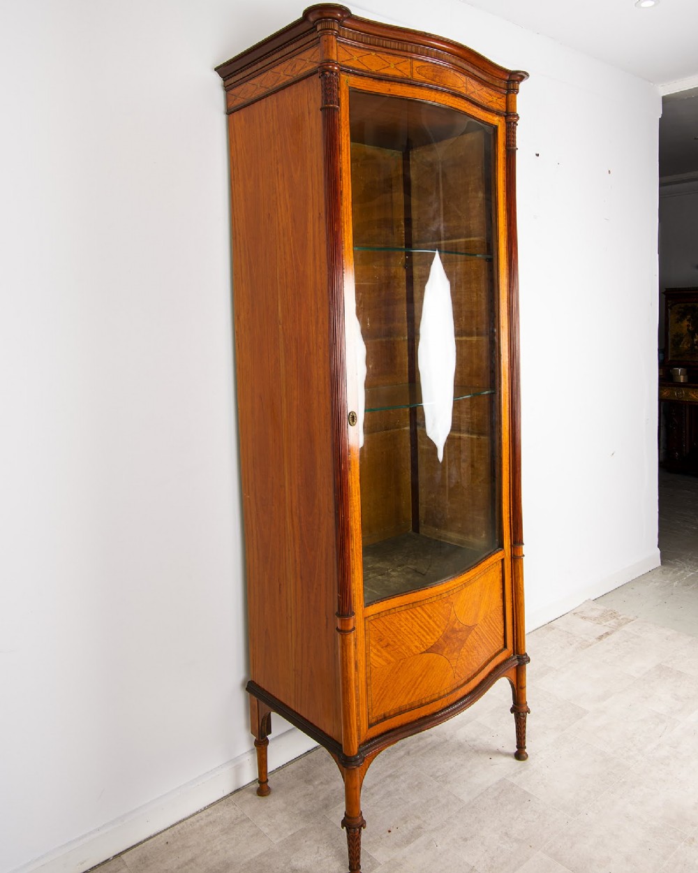 A tall Edwardian satinwood display cabinet featuring a curved glazed door, glass shelves, fluted corner columns, and geometric marquetry inlay. View VI