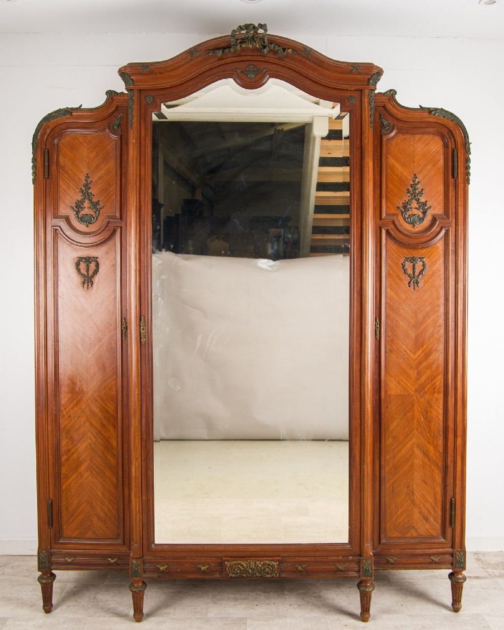 An antique French mahogany three-door armoire featuring a central arched mirrored door flanked by parquetry wood panels with gilt bronze ormolu mounts.
