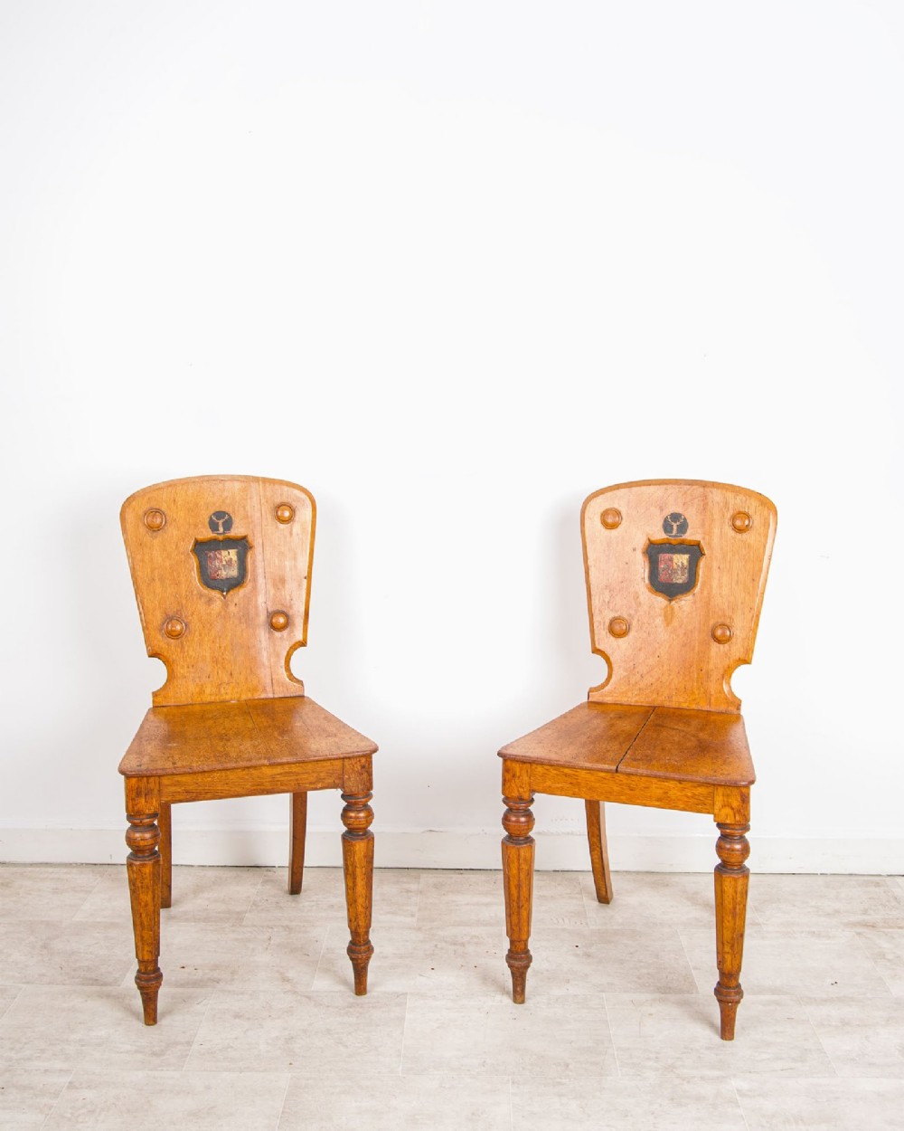 A pair of antique oak hall chairs featuring solid timber seats, turned front legs, and backrests decorated with carved bosses and painted heraldic shields.