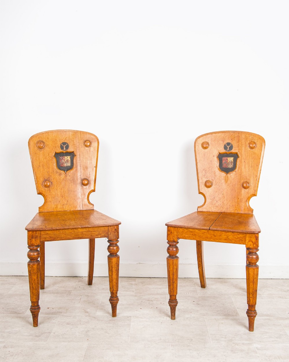 A pair of antique oak hall chairs featuring solid timber seats, turned front legs, and backrests decorated with carved bosses and painted heraldic shields. View II