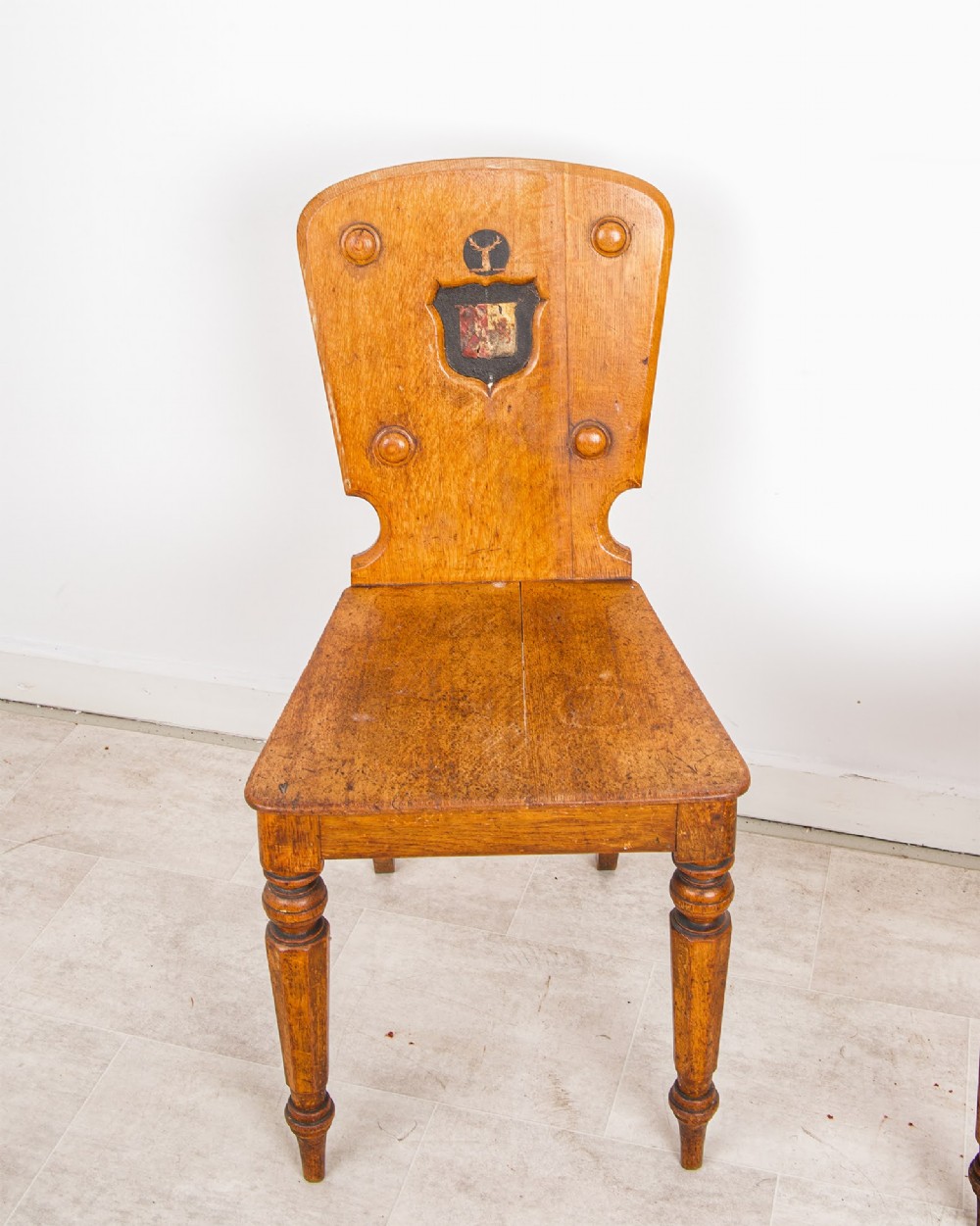A pair of antique oak hall chairs featuring solid timber seats, turned front legs, and backrests decorated with carved bosses and painted heraldic shields. View III