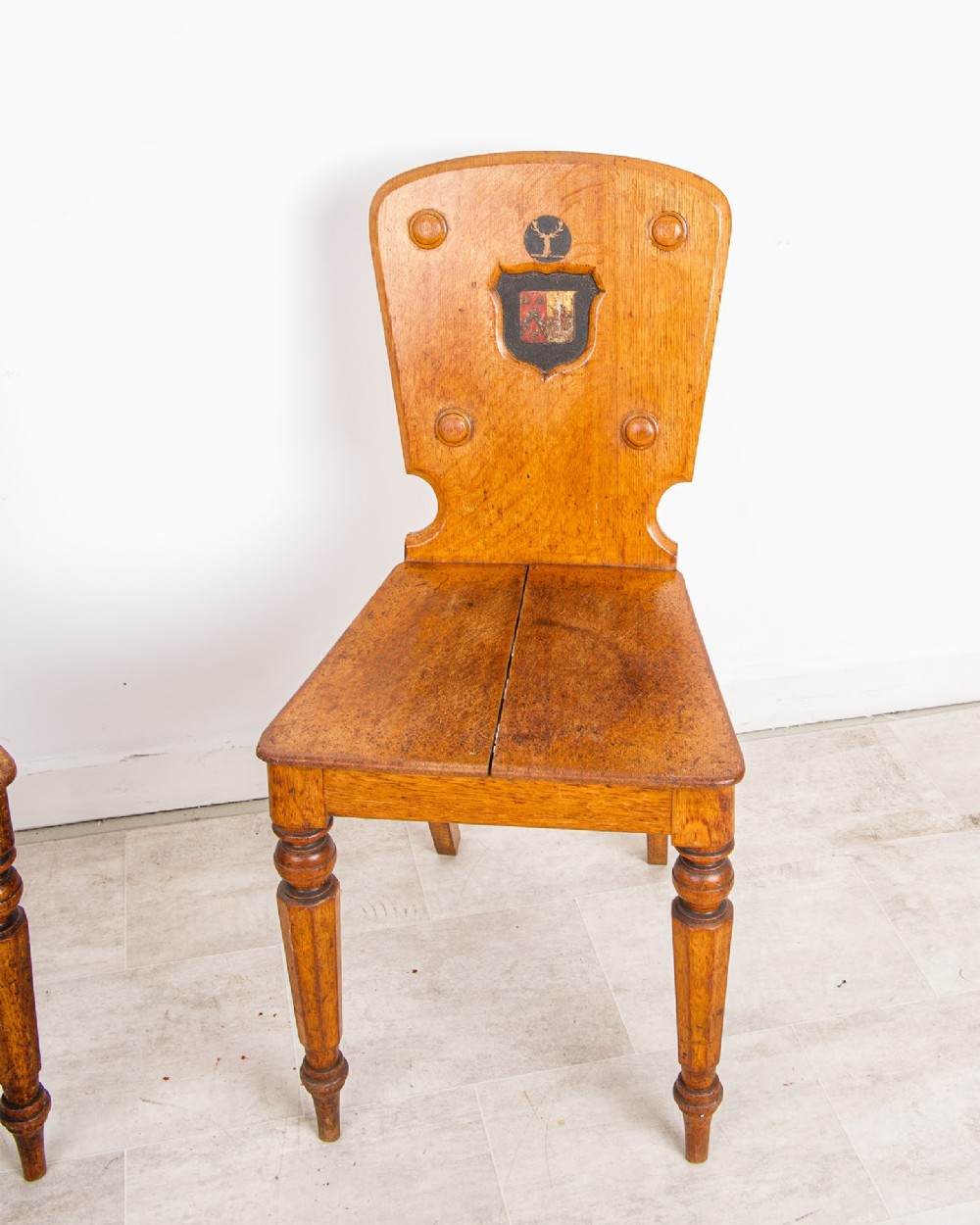 A pair of antique oak hall chairs featuring solid timber seats, turned front legs, and backrests decorated with carved bosses and painted heraldic shields. View IV