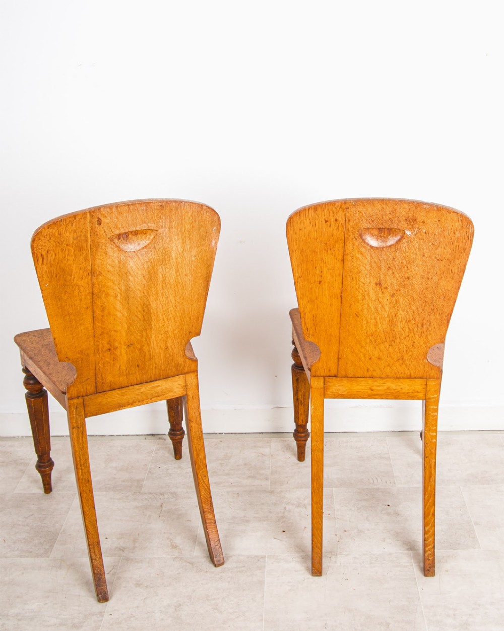 A pair of antique oak hall chairs featuring solid timber seats, turned front legs, and backrests decorated with carved bosses and painted heraldic shields. View V