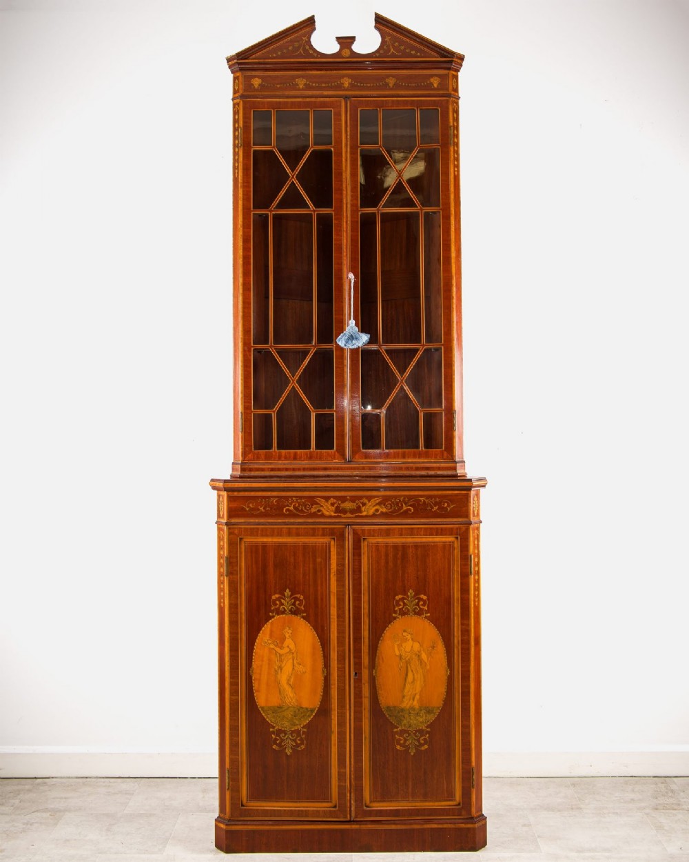 A tall Edwardian mahogany corner display cabinet featuring astragal glazed doors, a broken pediment, and classical inlaid oval panels on the lower doors.