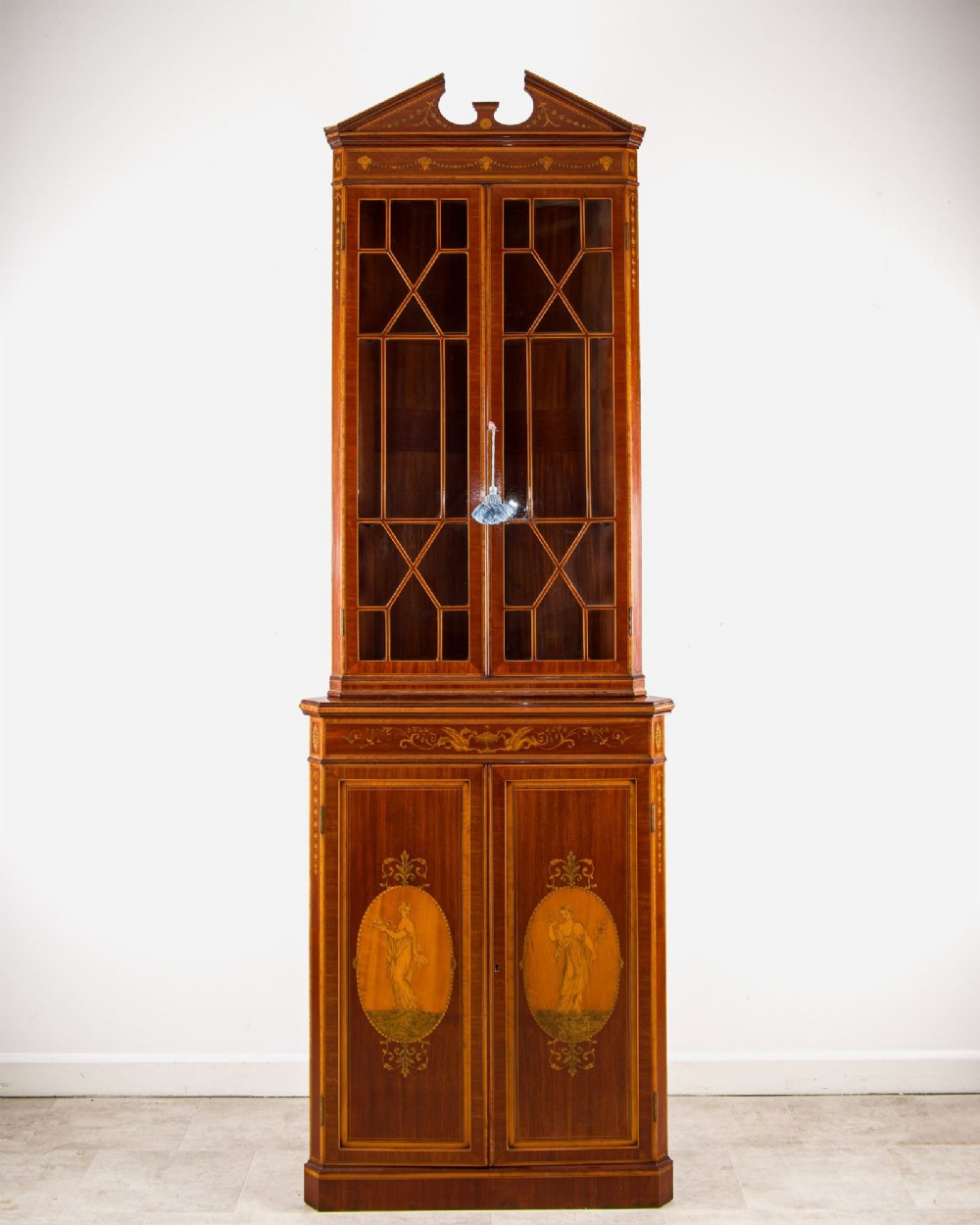 A tall Edwardian mahogany corner display cabinet featuring astragal glazed doors, a broken pediment, and classical inlaid oval panels on the lower doors. View II