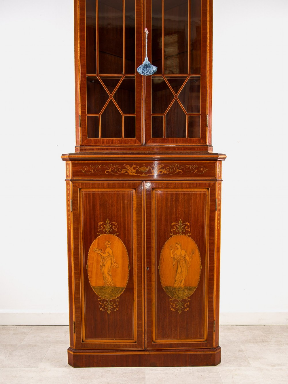 A tall Edwardian mahogany corner display cabinet featuring astragal glazed doors, a broken pediment, and classical inlaid oval panels on the lower doors. View IV