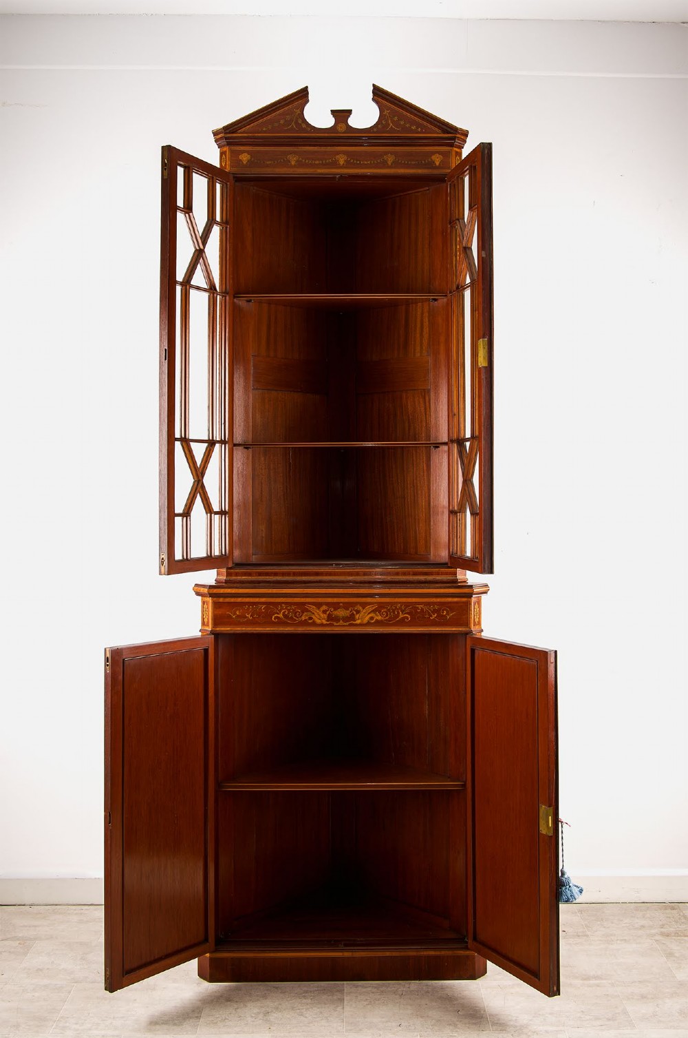 A tall Edwardian mahogany corner display cabinet featuring astragal glazed doors, a broken pediment, and classical inlaid oval panels on the lower doors. View VI