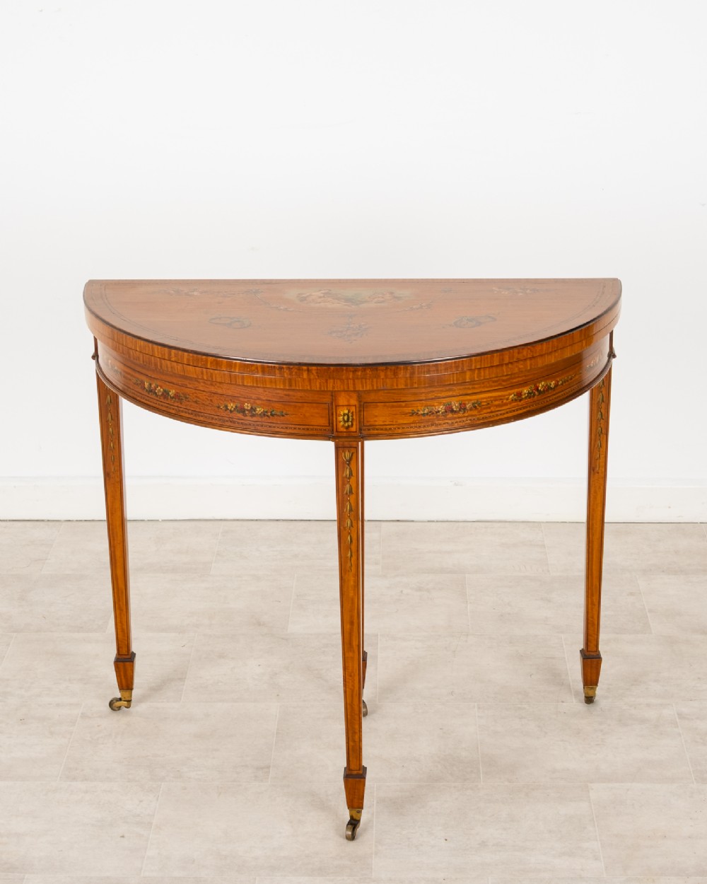 An Edwardian satinwood demi-lune games table featuring painted floral details and tapered legs on brass castors.