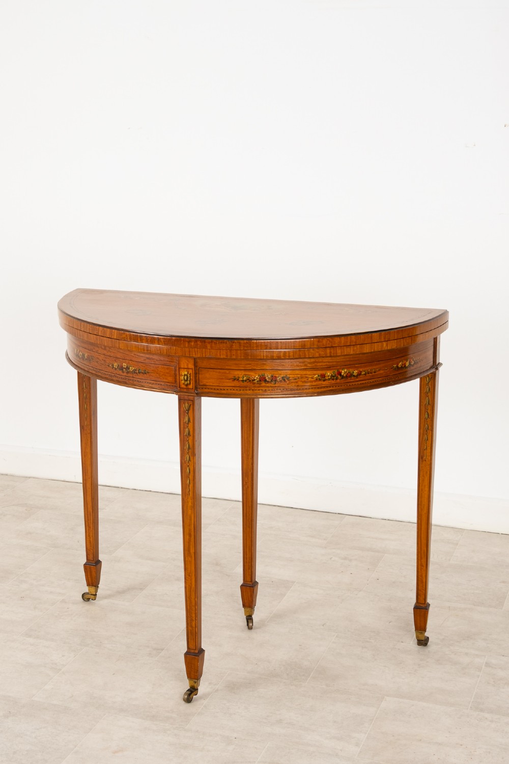 An Edwardian satinwood demi-lune games table featuring painted floral details and tapered legs on brass castors. View II