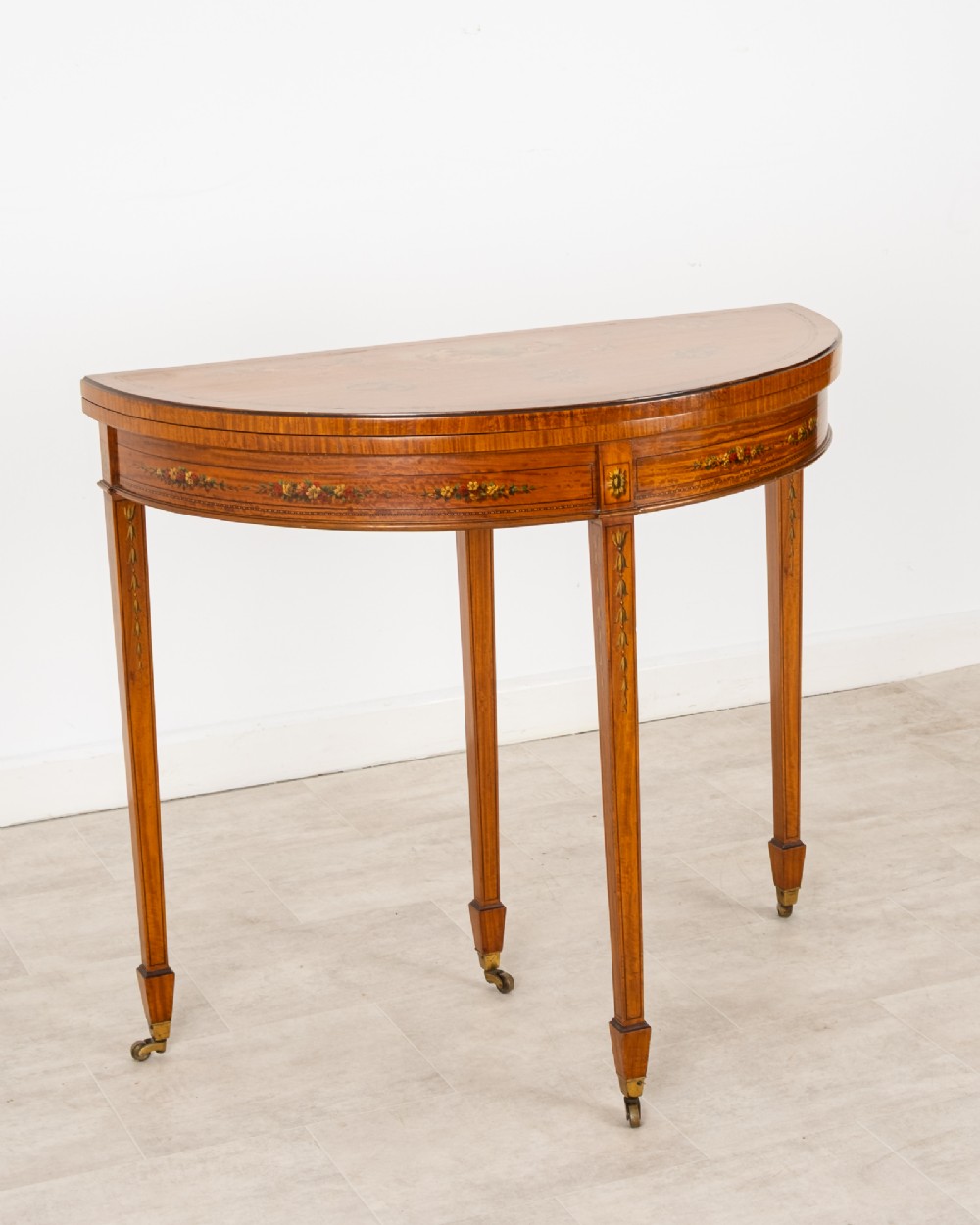 An Edwardian satinwood demi-lune games table featuring painted floral details and tapered legs on brass castors. View III