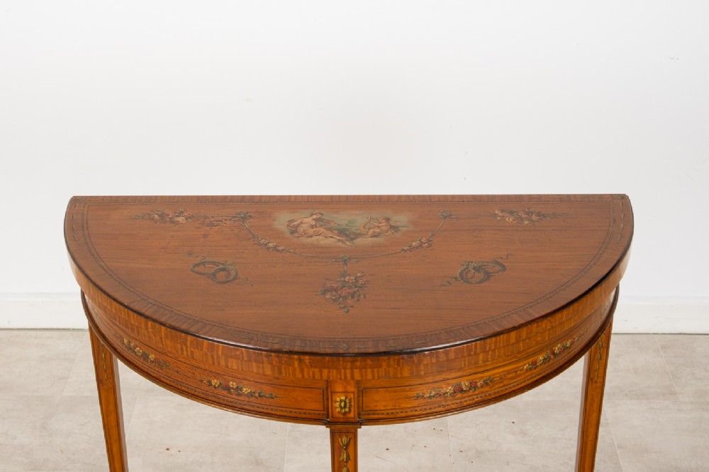 An Edwardian satinwood demi-lune games table featuring painted floral details and tapered legs on brass castors. View IV