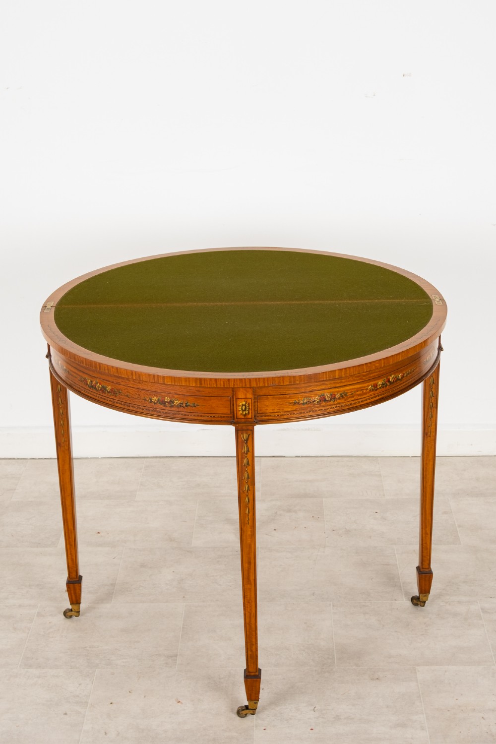 An Edwardian satinwood demi-lune games table featuring painted floral details and tapered legs on brass castors. View V