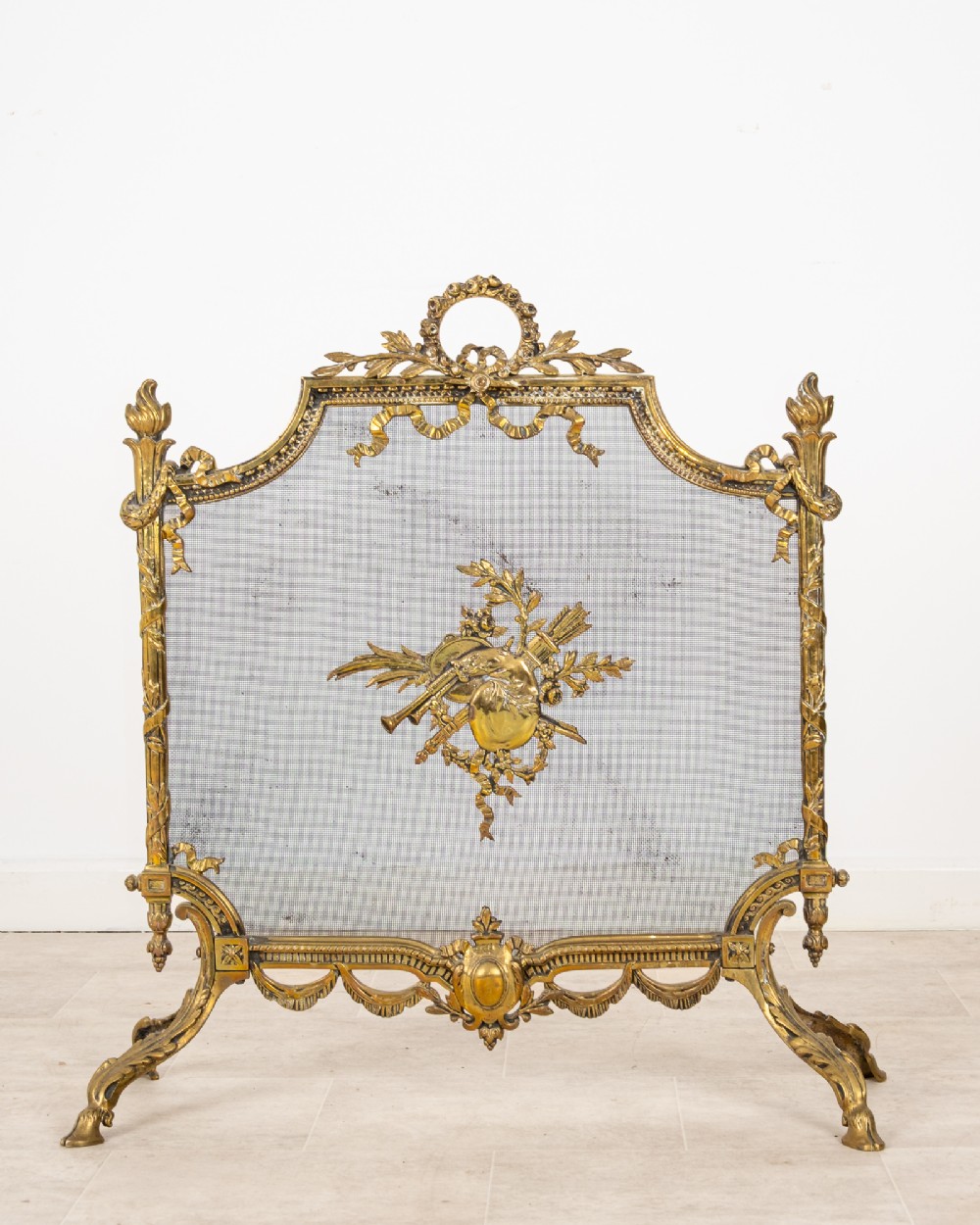 An antique French Louis XVI style gilt brass fire screen featuring a woven mesh panel decorated with a central trophy motif, ribbon bow cresting, and scrolled feet.