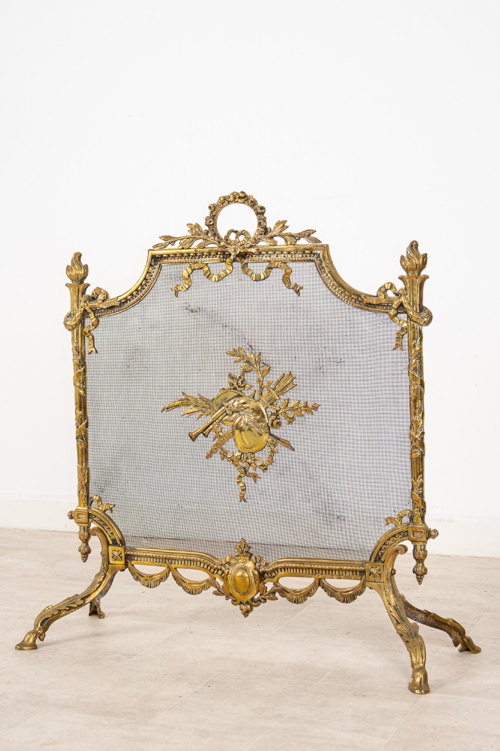 An antique French Louis XVI style gilt brass fire screen featuring a woven mesh panel decorated with a central trophy motif, ribbon bow cresting, and scrolled feet. View II