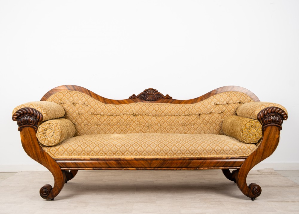A Victorian rosewood settee featuring carved scroll ends, a central carved cresting, and patterned mustard yellow upholstery with bolster cushions.