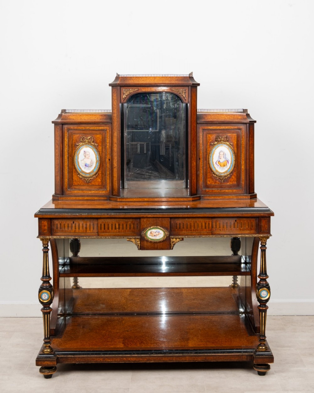 An antique French amboyna and ebonised bonheur du jour writing desk featuring inset porcelain plaques, gilt metal mounts, a central mirrored superstructure, and lower tier shelving.