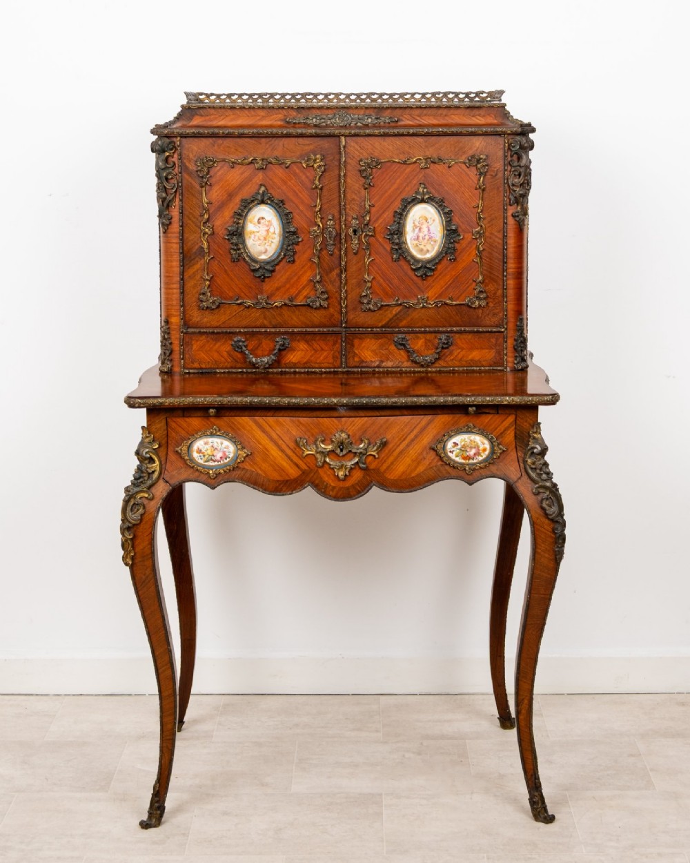 An antique French kingwood bonheur du jour writing desk featuring an upper cabinet with porcelain plaques, ornate ormolu mounts, and slender cabriole legs.