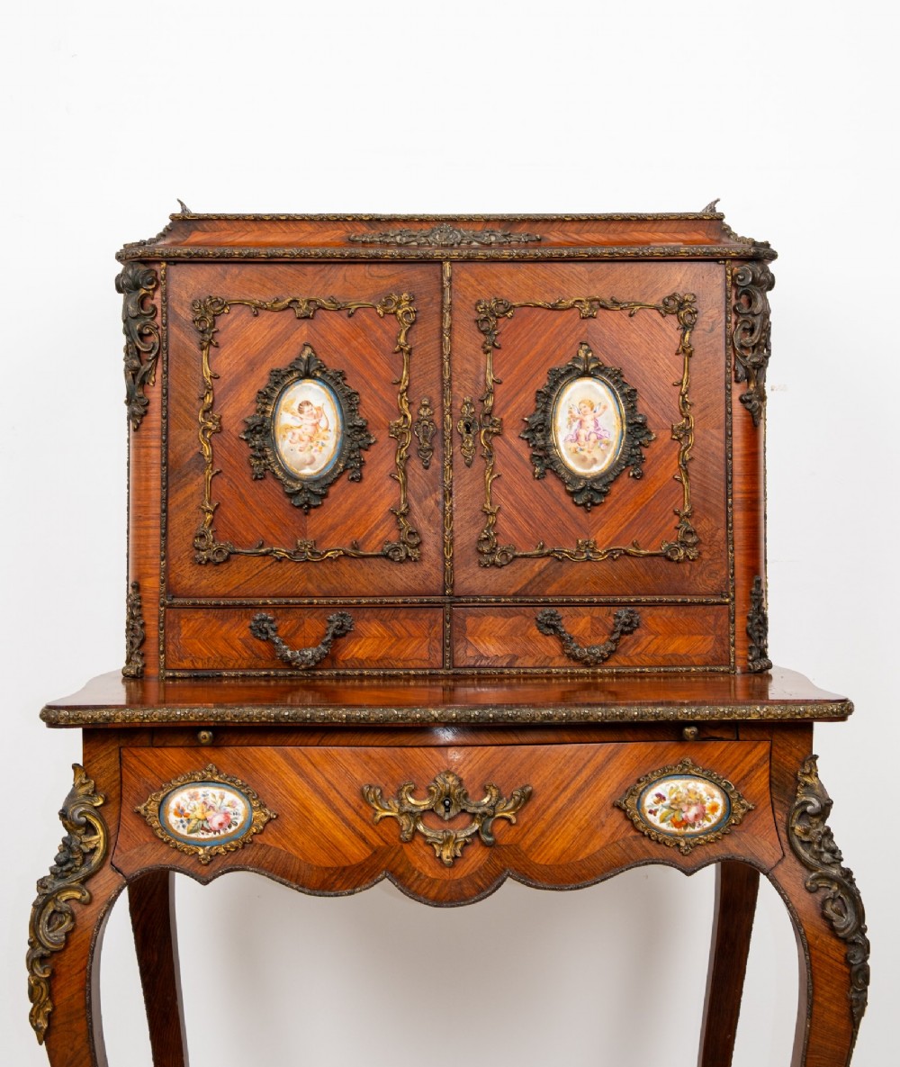 An antique French kingwood bonheur du jour writing desk featuring an upper cabinet with porcelain plaques, ornate ormolu mounts, and slender cabriole legs. View II
