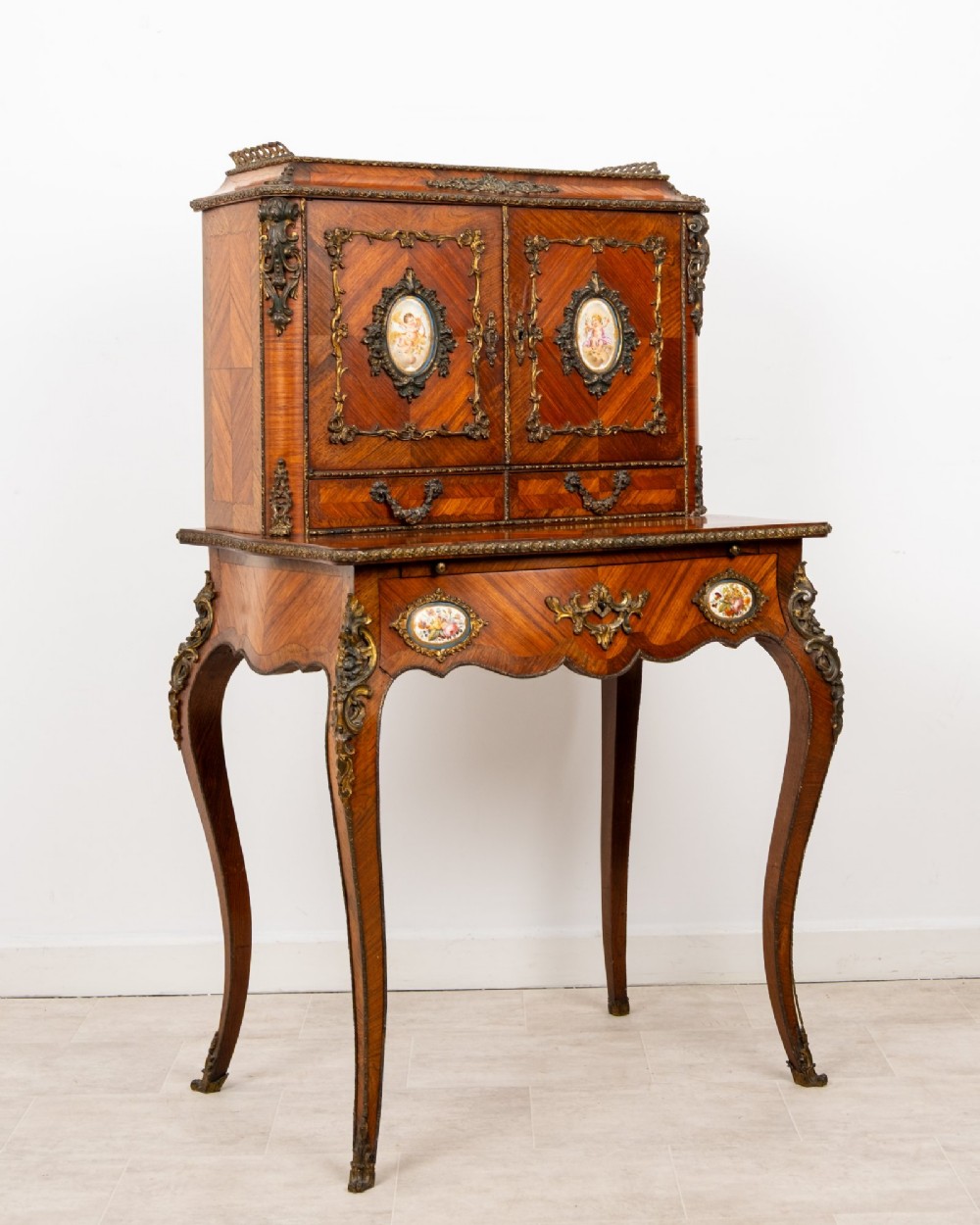 An antique French kingwood bonheur du jour writing desk featuring an upper cabinet with porcelain plaques, ornate ormolu mounts, and slender cabriole legs. View III