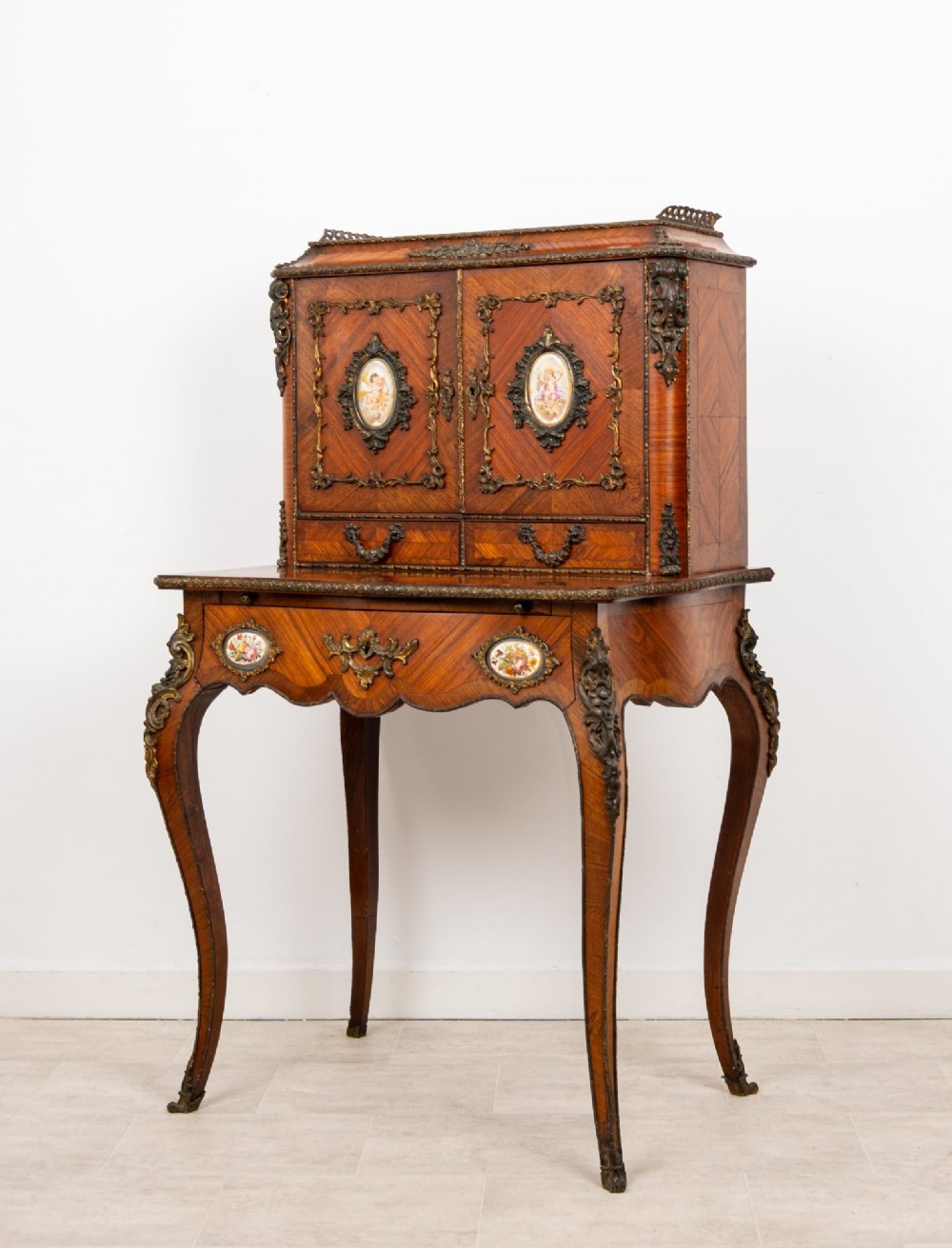 An antique French kingwood bonheur du jour writing desk featuring an upper cabinet with porcelain plaques, ornate ormolu mounts, and slender cabriole legs. View IV