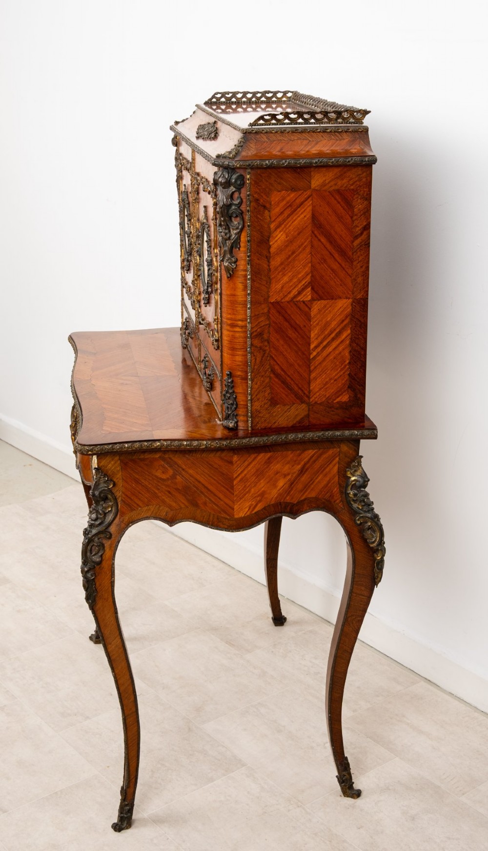 An antique French kingwood bonheur du jour writing desk featuring an upper cabinet with porcelain plaques, ornate ormolu mounts, and slender cabriole legs. View V
