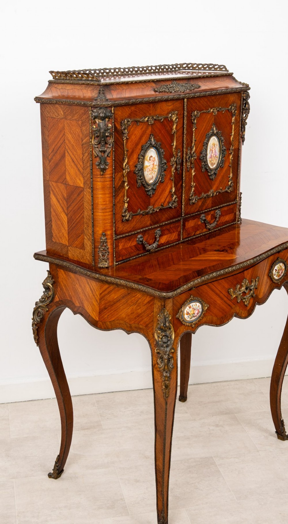 An antique French kingwood bonheur du jour writing desk featuring an upper cabinet with porcelain plaques, ornate ormolu mounts, and slender cabriole legs. View VI