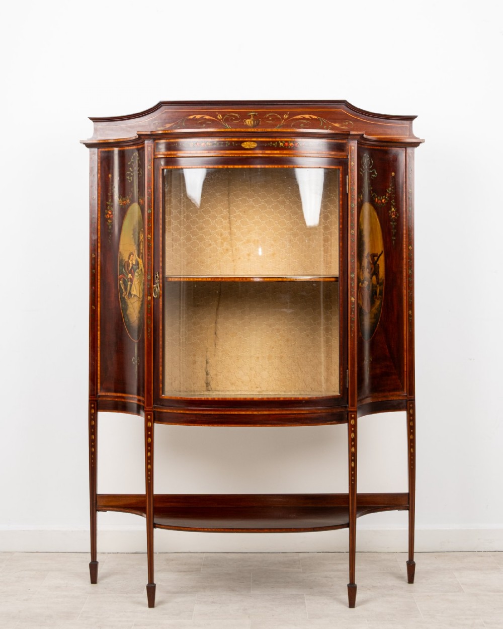An Edwardian mahogany bow-front display cabinet featuring hand-painted classical scene panels, satinwood inlay, glass door, and tapered spade legs.