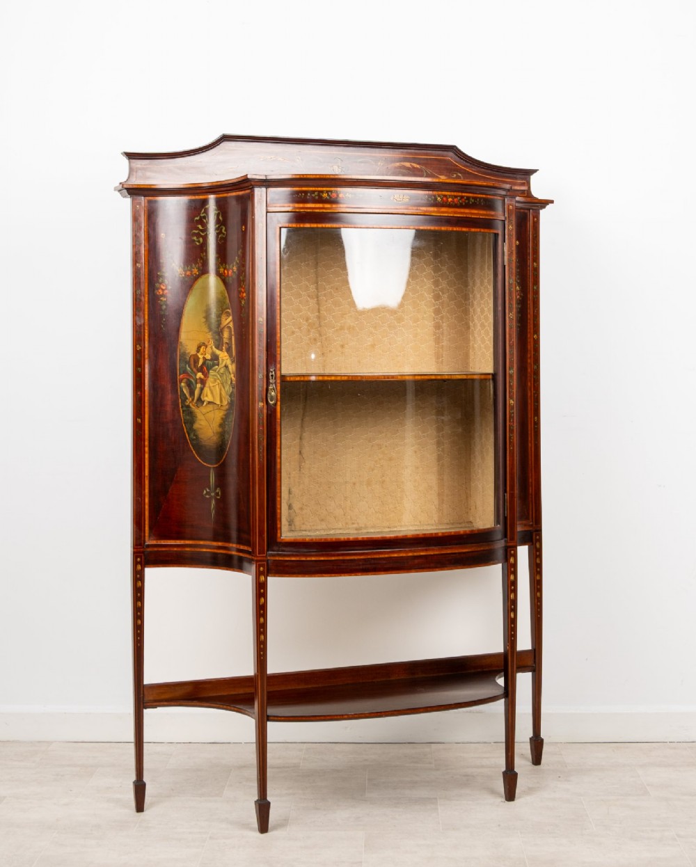 An Edwardian mahogany bow-front display cabinet featuring hand-painted classical scene panels, satinwood inlay, glass door, and tapered spade legs. View II