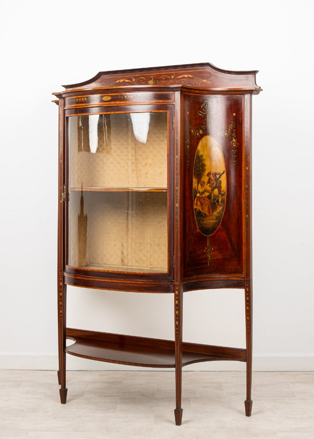 An Edwardian mahogany bow-front display cabinet featuring hand-painted classical scene panels, satinwood inlay, glass door, and tapered spade legs. View III