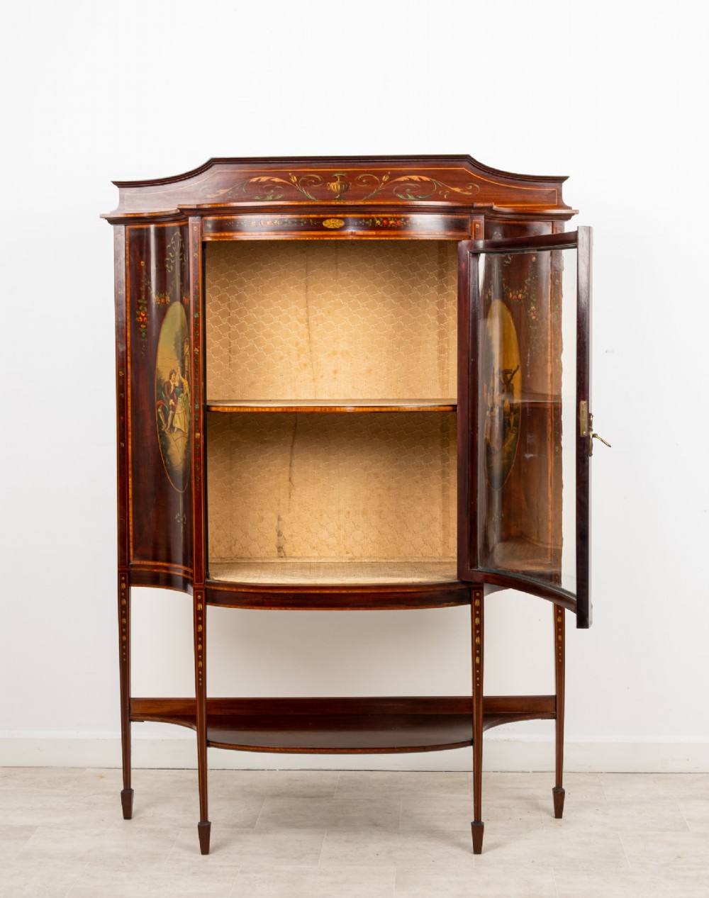 An Edwardian mahogany bow-front display cabinet featuring hand-painted classical scene panels, satinwood inlay, glass door, and tapered spade legs. View V