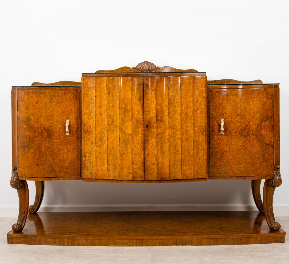 An Art Deco burr walnut sideboard featuring a stepped top with a fluted central door, curved end cupboards, decorative handles, and carved legs resting on a continuous plinth.