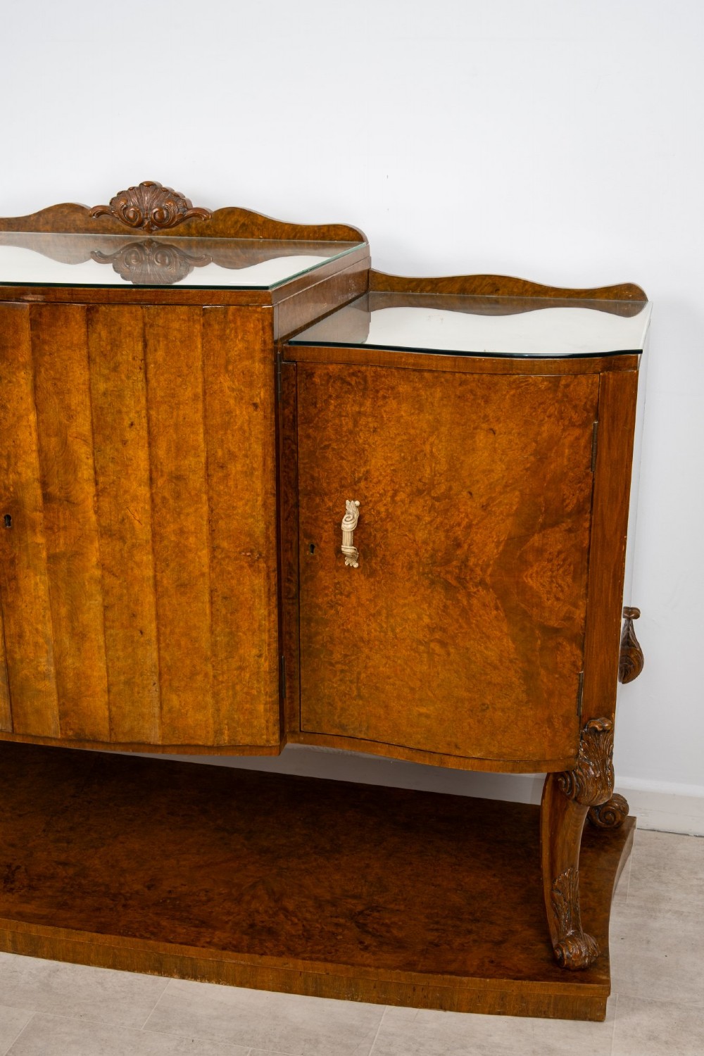An Art Deco burr walnut sideboard featuring a stepped top with a fluted central door, curved end cupboards, decorative handles, and carved legs resting on a continuous plinth. View IV