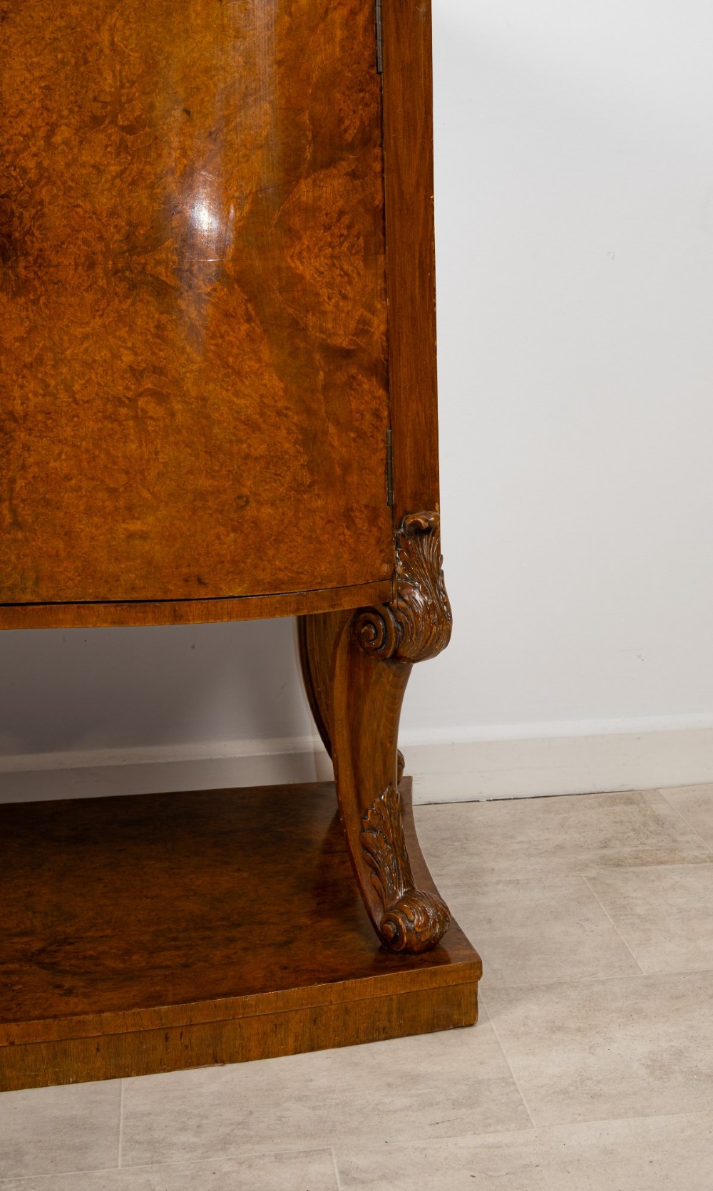 An Art Deco burr walnut sideboard featuring a stepped top with a fluted central door, curved end cupboards, decorative handles, and carved legs resting on a continuous plinth. View V