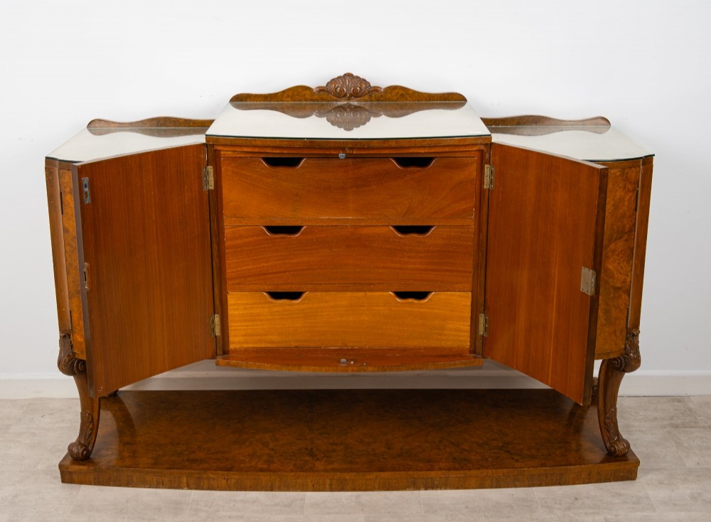 An Art Deco burr walnut sideboard featuring a stepped top with a fluted central door, curved end cupboards, decorative handles, and carved legs resting on a continuous plinth. View VI