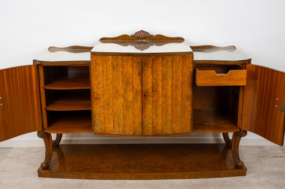 An Art Deco burr walnut sideboard featuring a stepped top with a fluted central door, curved end cupboards, decorative handles, and carved legs resting on a continuous plinth. View VII