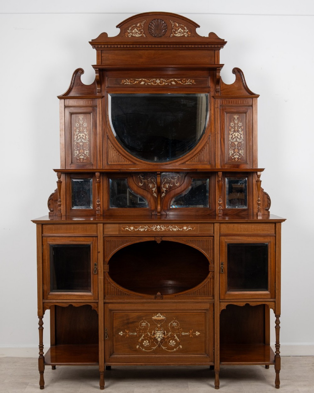 A late Victorian mahogany chiffonier cabinet featuring floral marquetry inlay, glass cupboard doors, open lower shelving, and an upper oval mirror back with a carved shell crest.