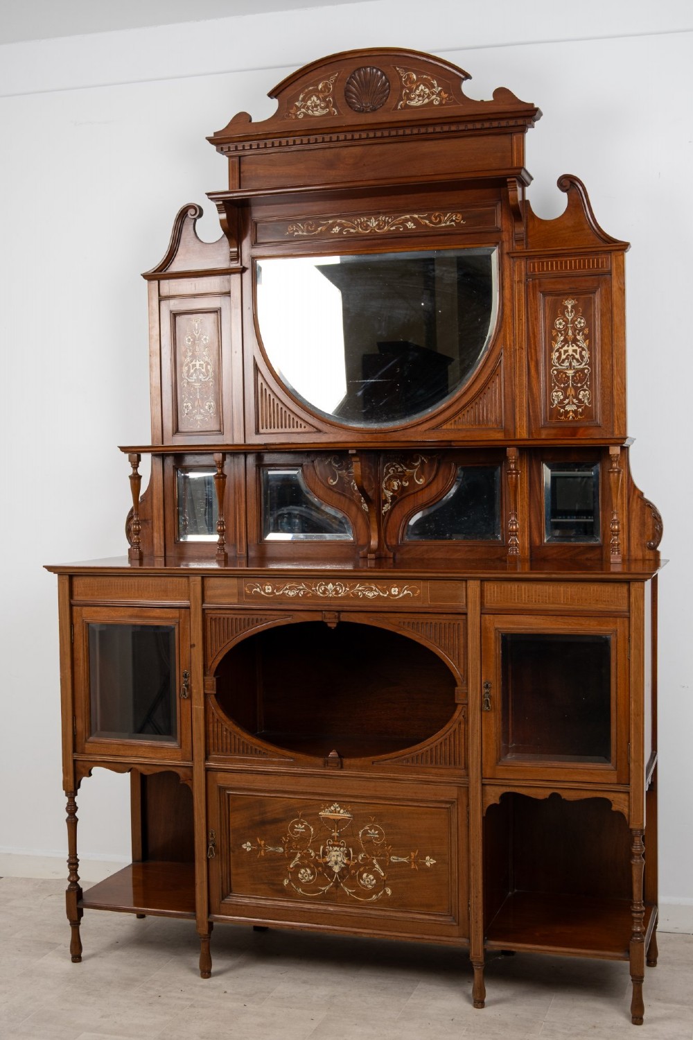 A late Victorian mahogany chiffonier cabinet featuring floral marquetry inlay, glass cupboard doors, open lower shelving, and an upper oval mirror back with a carved shell crest. View II
