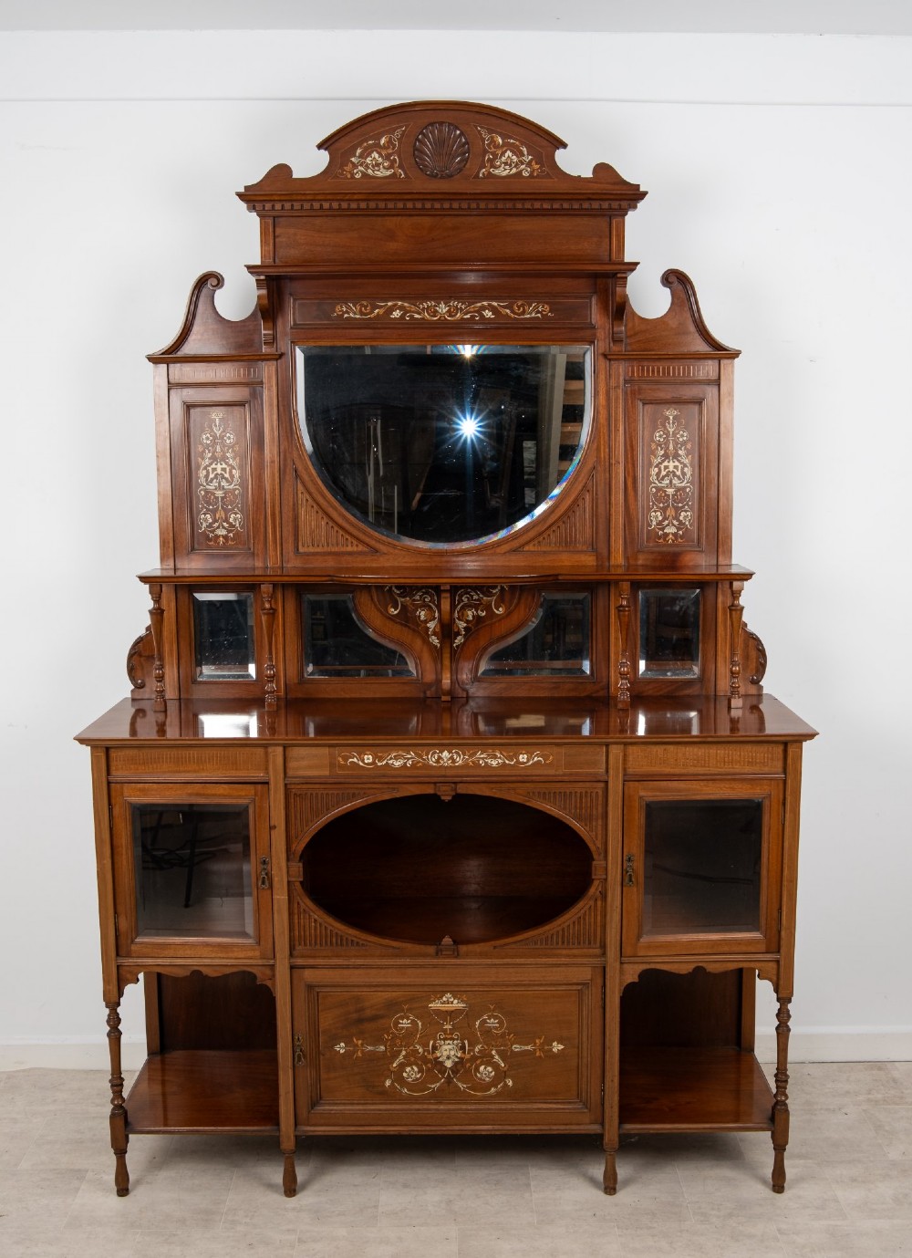 A late Victorian mahogany chiffonier cabinet featuring floral marquetry inlay, glass cupboard doors, open lower shelving, and an upper oval mirror back with a carved shell crest. View IV