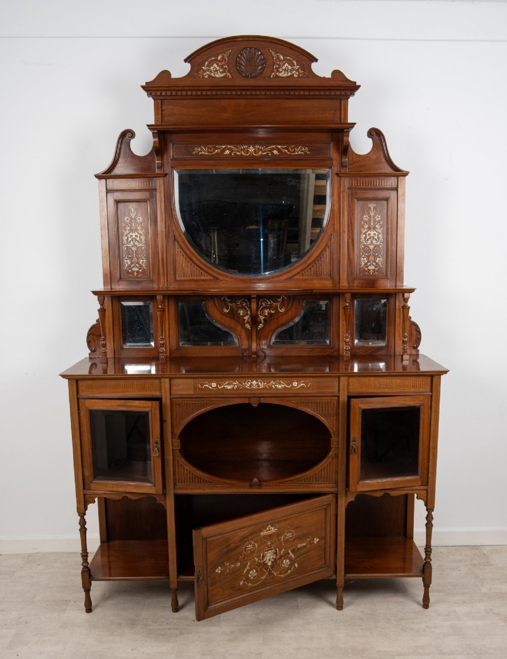 A late Victorian mahogany chiffonier cabinet featuring floral marquetry inlay, glass cupboard doors, open lower shelving, and an upper oval mirror back with a carved shell crest. View X