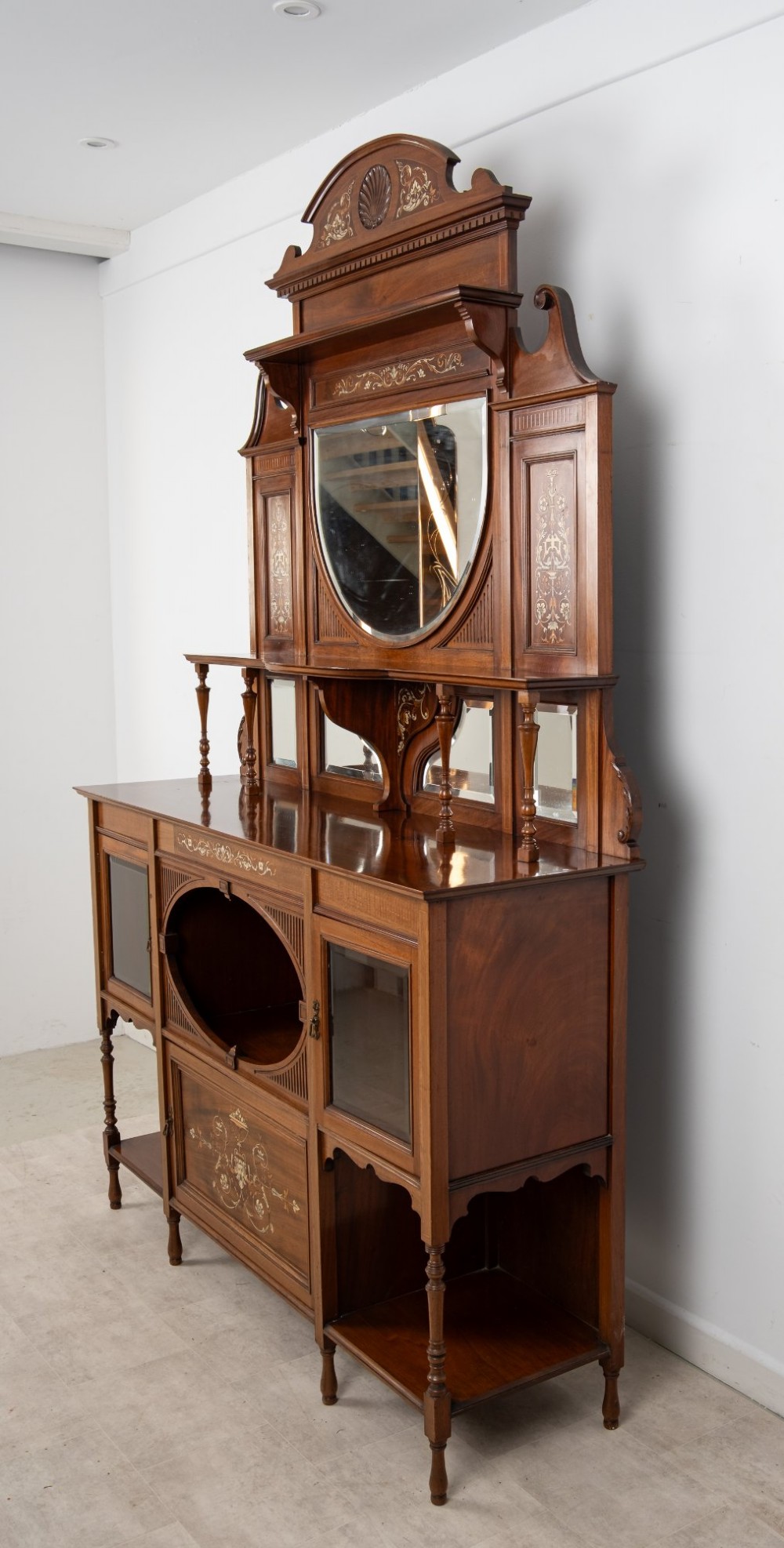 A late Victorian mahogany chiffonier cabinet featuring floral marquetry inlay, glass cupboard doors, open lower shelving, and an upper oval mirror back with a carved shell crest. View XI