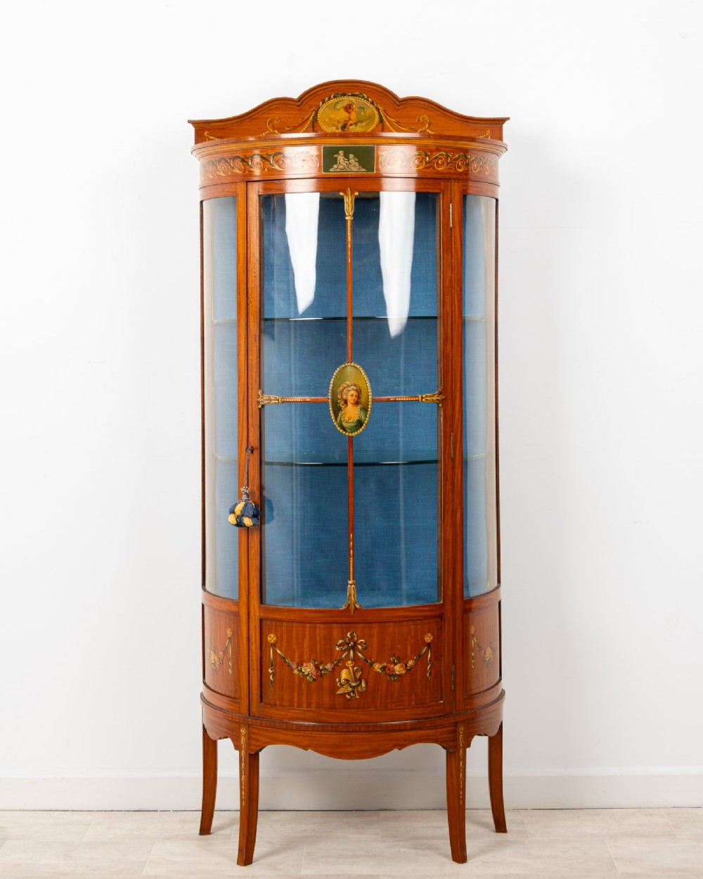 An Edwardian satinwood bow-front vitrine display cabinet featuring hand-painted floral swags, a central painted portrait medallion, curved glass panels, and a blue fabric-lined interior.