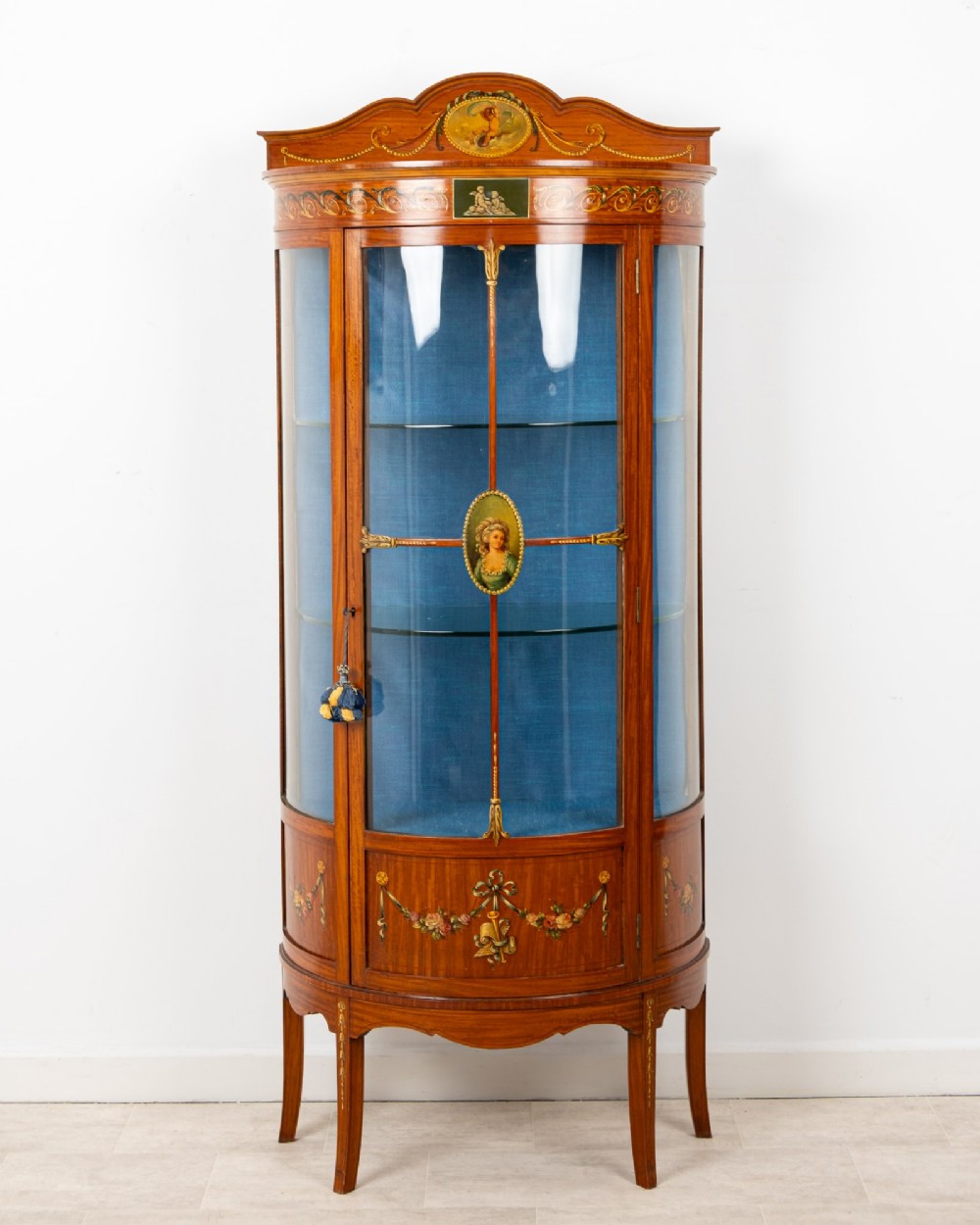 An Edwardian satinwood bow-front vitrine display cabinet featuring hand-painted floral swags, a central painted portrait medallion, curved glass panels, and a blue fabric-lined interior. View II