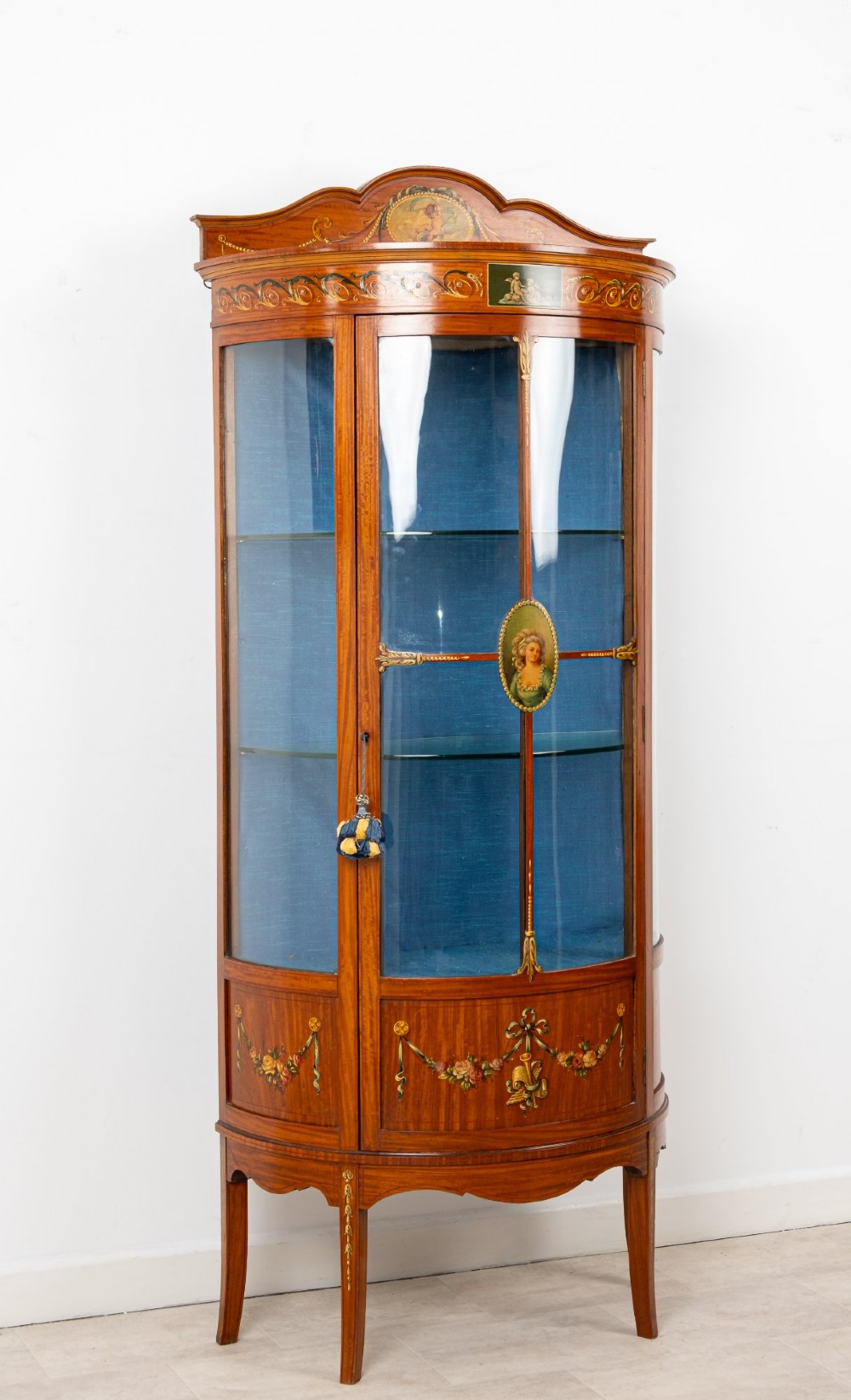 An Edwardian satinwood bow-front vitrine display cabinet featuring hand-painted floral swags, a central painted portrait medallion, curved glass panels, and a blue fabric-lined interior. View IV