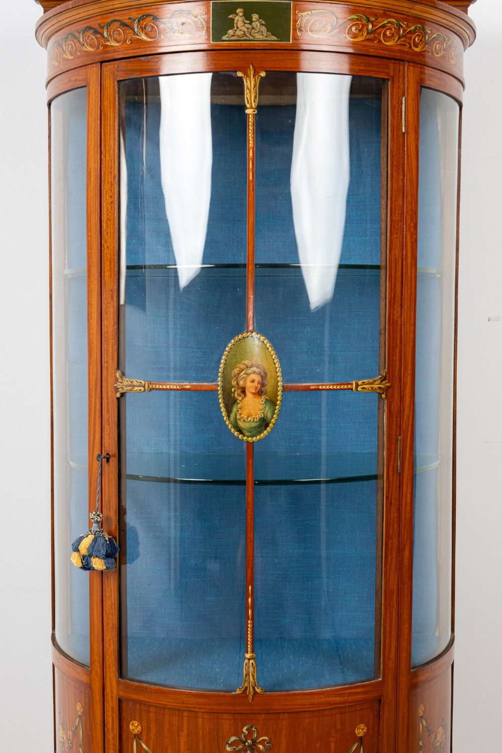 An Edwardian satinwood bow-front vitrine display cabinet featuring hand-painted floral swags, a central painted portrait medallion, curved glass panels, and a blue fabric-lined interior. View V