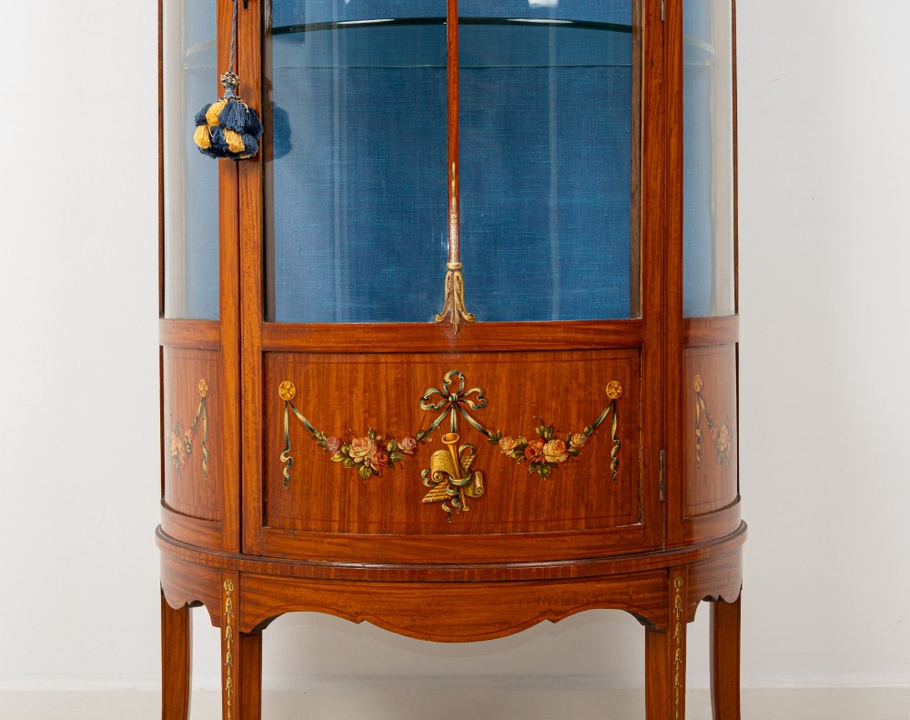 An Edwardian satinwood bow-front vitrine display cabinet featuring hand-painted floral swags, a central painted portrait medallion, curved glass panels, and a blue fabric-lined interior. View VI