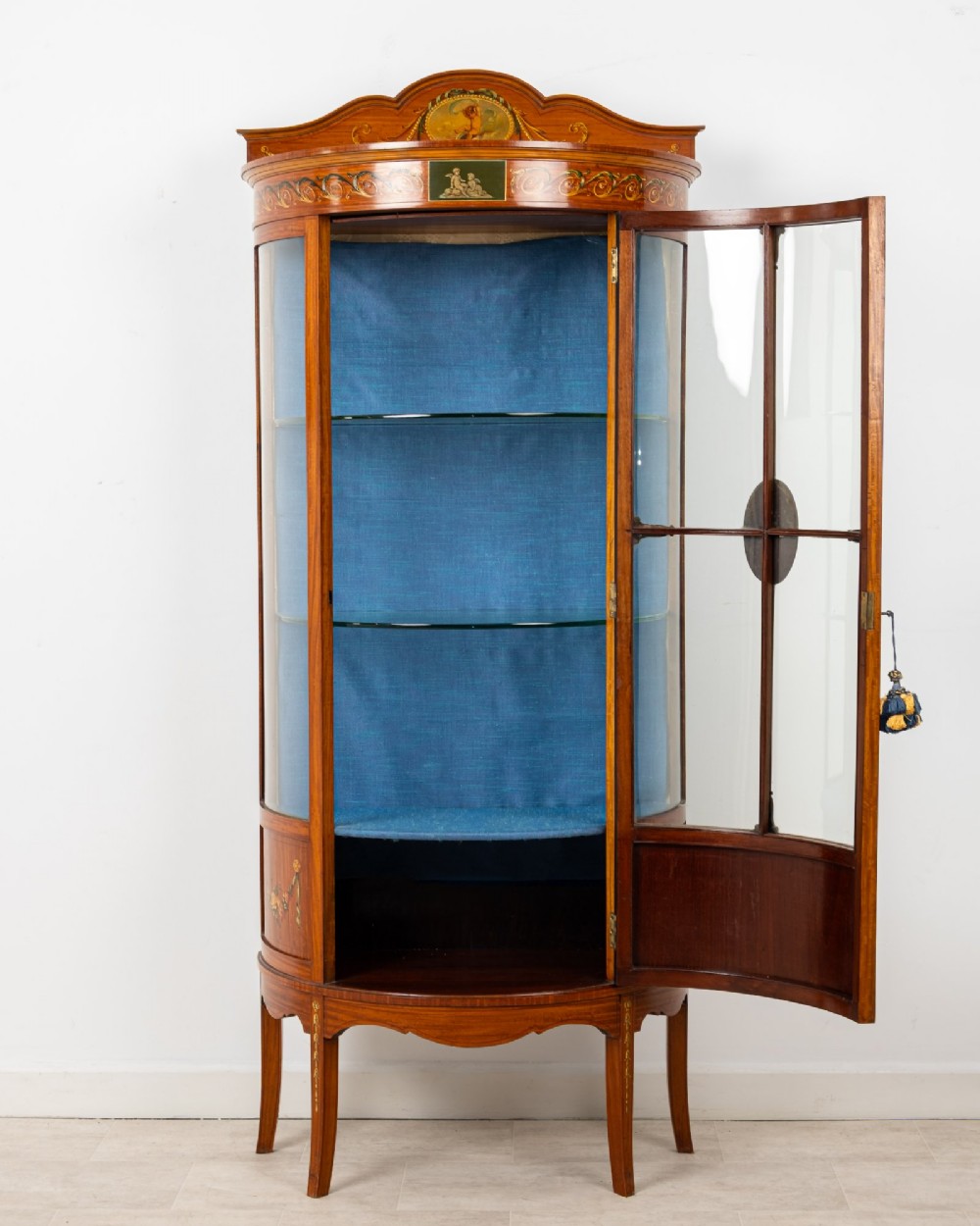 An Edwardian satinwood bow-front vitrine display cabinet featuring hand-painted floral swags, a central painted portrait medallion, curved glass panels, and a blue fabric-lined interior. View IX