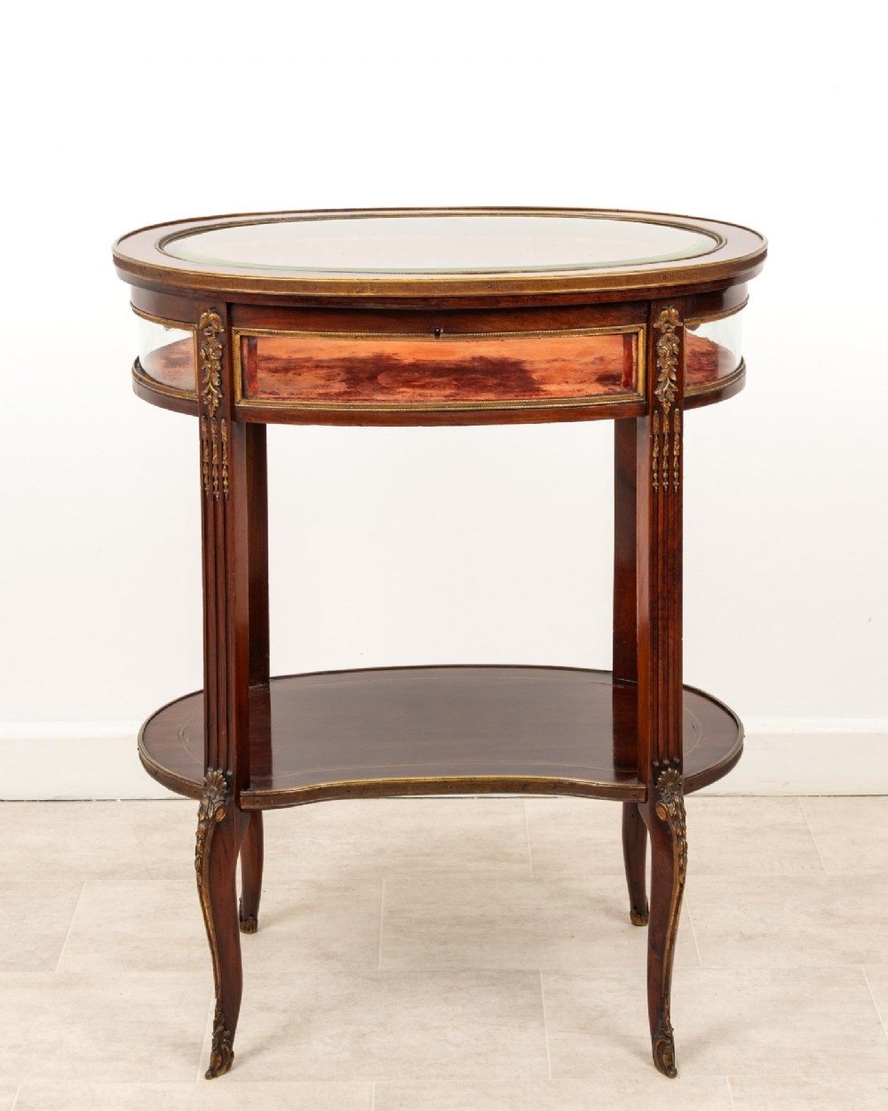 An antique oval mahogany bijouterie table featuring curved glass sides, an undertier shelf, and ornate gilt-bronze mounts.