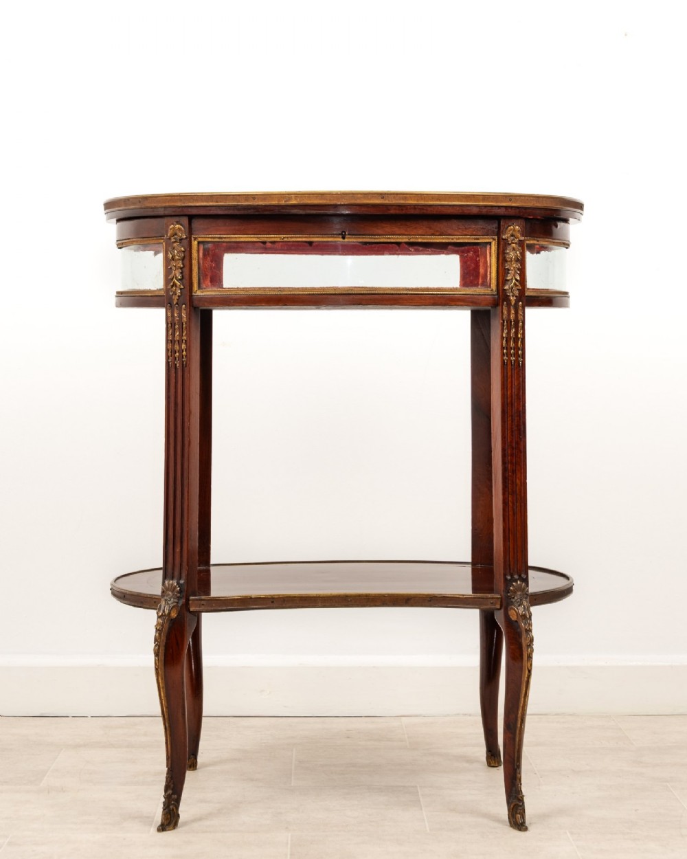 An antique oval mahogany bijouterie table featuring curved glass sides, an undertier shelf, and ornate gilt-bronze mounts. View II